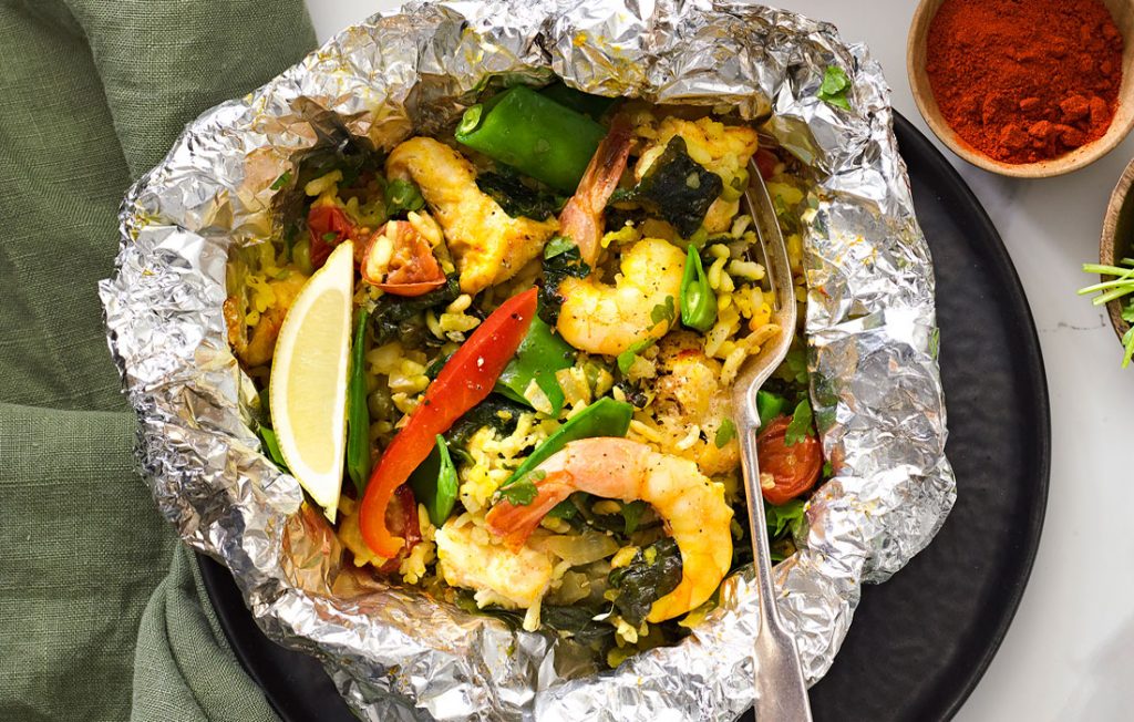 Barbecue seafood packet paella Foil packet with rice, prawns vegetables, lemon and fresh herbs on a marble table top with a dish of paprika and fresh parsley.