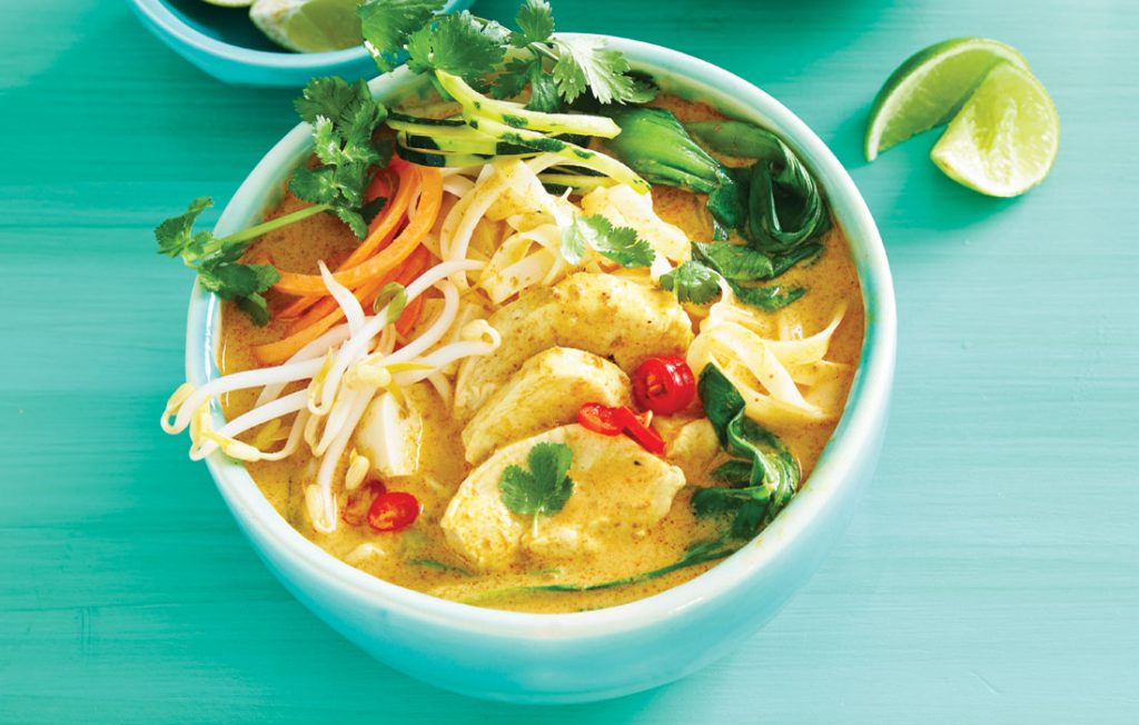 Chicken and vegetable laksa Bowl of laksa soup with chicken, noodles, mung bean sprouts, grated vegetables and fresh herbs. Lime wedges on the side. Green table top.