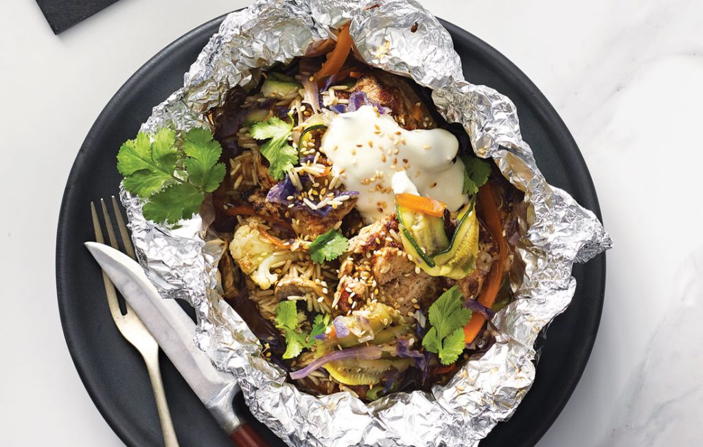 Five-spice chicken packets Foil packet with chicken, yohurt, vegetables, seeds, frsh herbs and rice on a black plate.