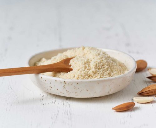 A bowl of almond meal with a wooden spoon and almonds scattered around